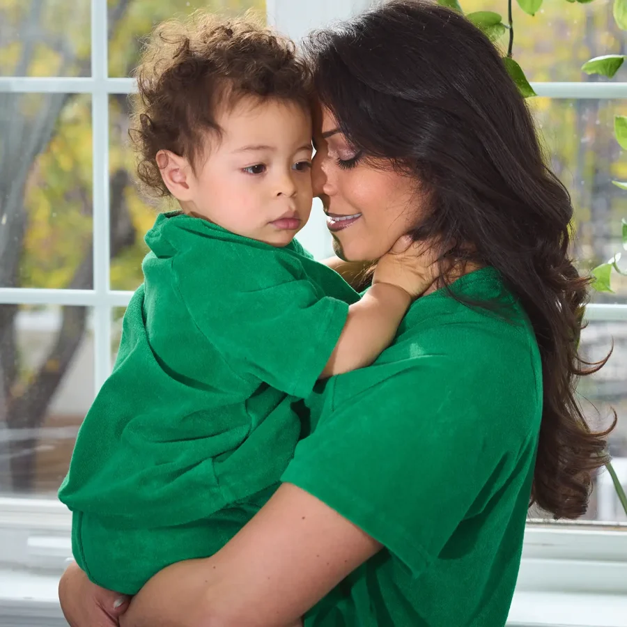 Mother and child wearing matching green terry cloth outfits, sharing a warm and loving embrace near a sunlit window.
